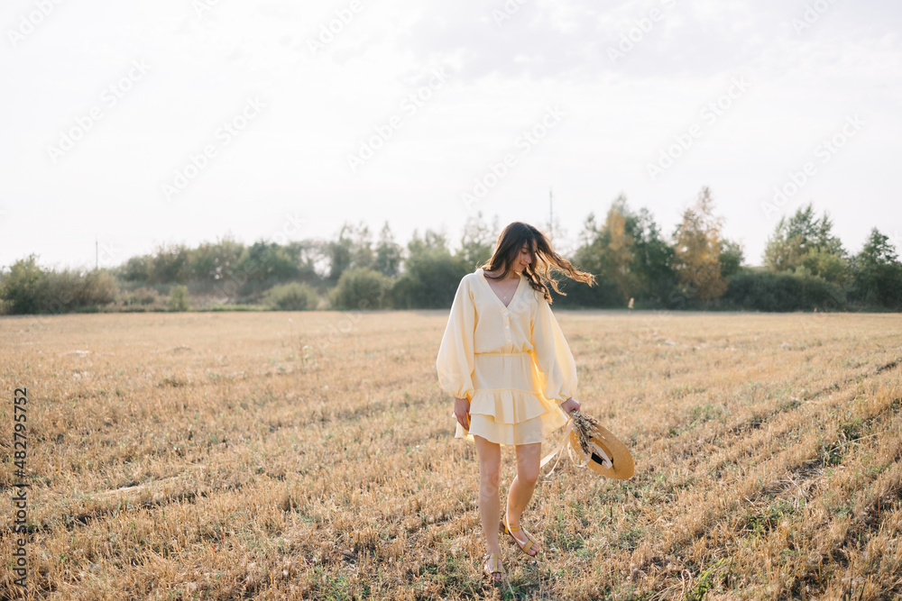 Caucasian young brunette walking through a wheat field. girl in yellow summer dress and straw hat. sheared golden ears of wheat, rye. Close-up portrait of a beautiful girl with brown eyes. 