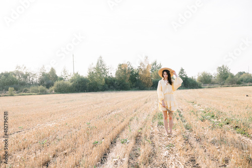 Young beautiful brunette on a summer day. girl in yellow dress. Large straw hat with a bouquet of wheat. Agricultural texture