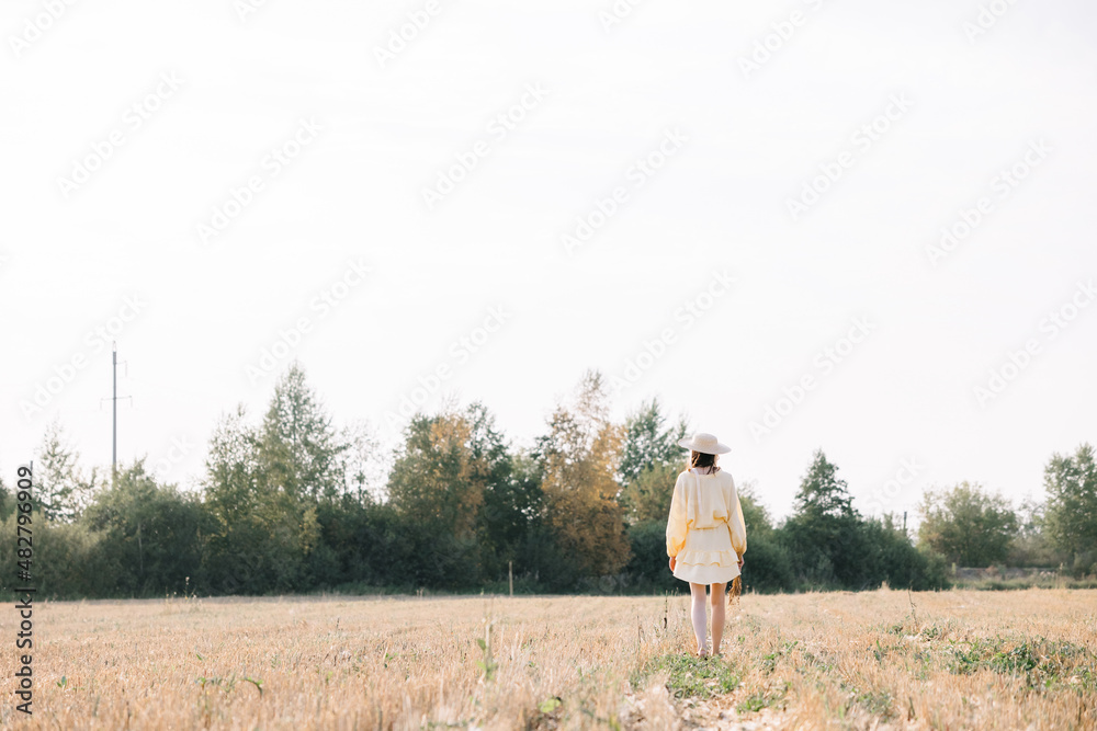 Obraz premium Young beautiful brunette on a summer day. girl in yellow dress. Large straw hat with a bouquet of wheat. Agricultural texture