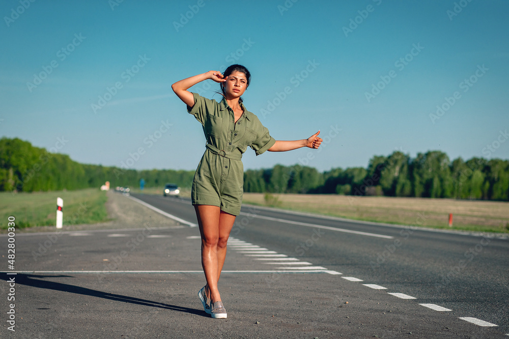 tanned pretty lady with long slender sexy legs in a tight overalls is trying to catch a car in the middle of the road, full height