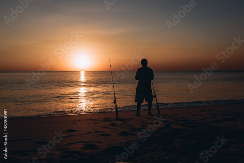 Silhouette of a man fishing on a background of beautiful colorful sunset next to the pleasant sea on the sandy beach.