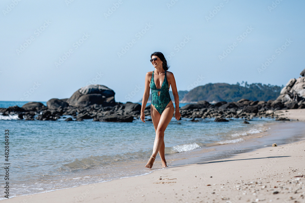 Wonderful young woman with beautiful long legs is walking on the beach with a statuesque gait on the background of rocks and stones