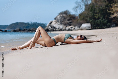 Attractive and young woman with a perfect body is lying on a wet beach throwing her arms up in the pleasure of the hot sun and sunbathing against the background of the rocky jungle and the sea.