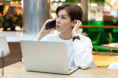 Young beautiful brunette woman sitting at a shopping center at a table and working at a computer laptop, using mobile phone. Freelance and business concept