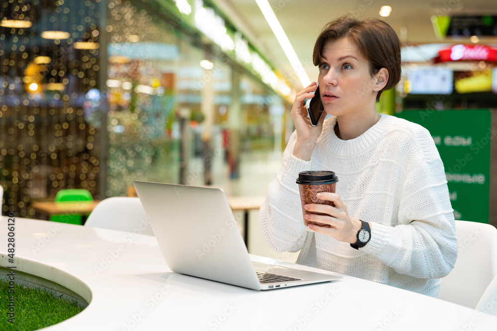 Young attractive woman sitting at a shopping center at a table and working at a computer laptop, using mobile phone, drinking coffee. Cafe interior. Freelance and business concept Young attractive woman sitting at a shopping center at a table and working at a computer laptop, using mobile phone, drinking coffee. Cafe interior. Freelance and business concept