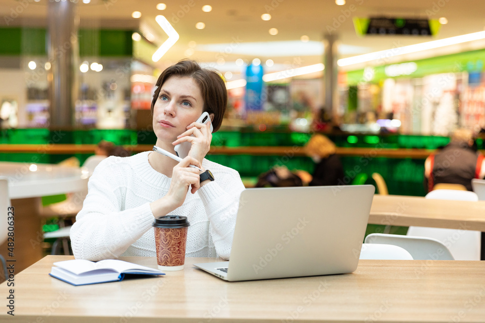 Young attractive brunette woman sitting at a shopping center at a table with coffee paper cup and working at a computer laptop, using mobile phone. Freelance and business concept