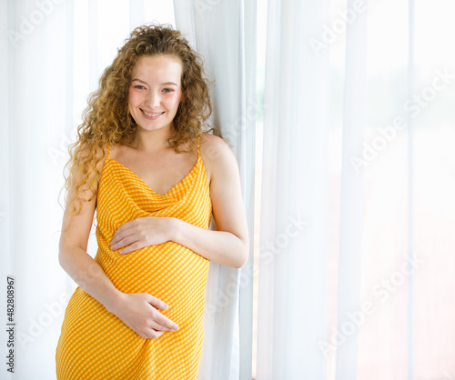 Curly hairstyle happy healthy Caucasian pregnancy mother model in orange stripe maternity long dress cloth standing smiling touching big belly look at camera in bedroom