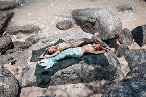 two sexy slim mermaids are sunbathing on stones on a fishnet, mythical creatures