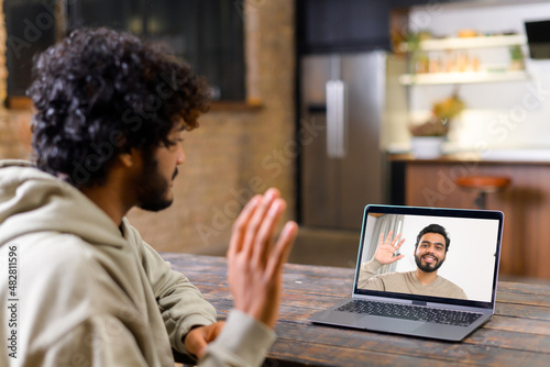 Cheerful attractive middle eastern guy is using laptop for video communication, while sitting at the table in modern flat and waving into webcam. Talking online concept