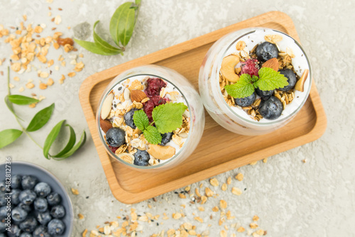 two glass glasses with a delicious healthy breakfast of granola with yogurt, nuts, berries and mint stand on a wooden tray. top view. gray background.
