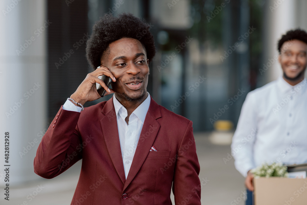 Portrait of handsome confident serious African American businessman CEO top manager in stylish maroon suit, talking on phone outside in the background of business center, company office Portrait of handsome confident serious African American businessman CEO top manager in stylish maroon suit, talking on phone outside in the background of business center, company office