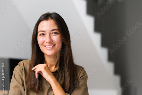 Portrait of charming elegant asian woman with hand under chin smiling at the camera, sitting in the modern house, wearing domestic clothes. Stock photo