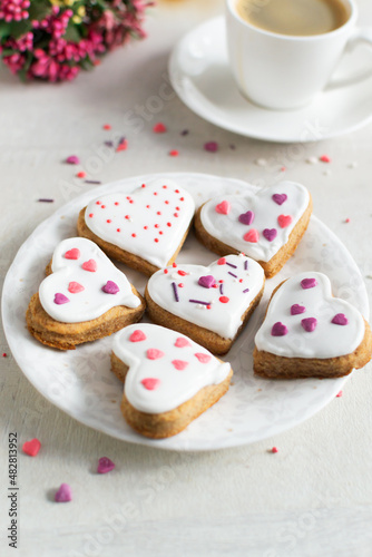 Gingerbread covered with white icing and sprinkling on a light saucer with a cup of espresso and flowers in the background. The concept of celebrating Valentine's Day. Vertical orientation.