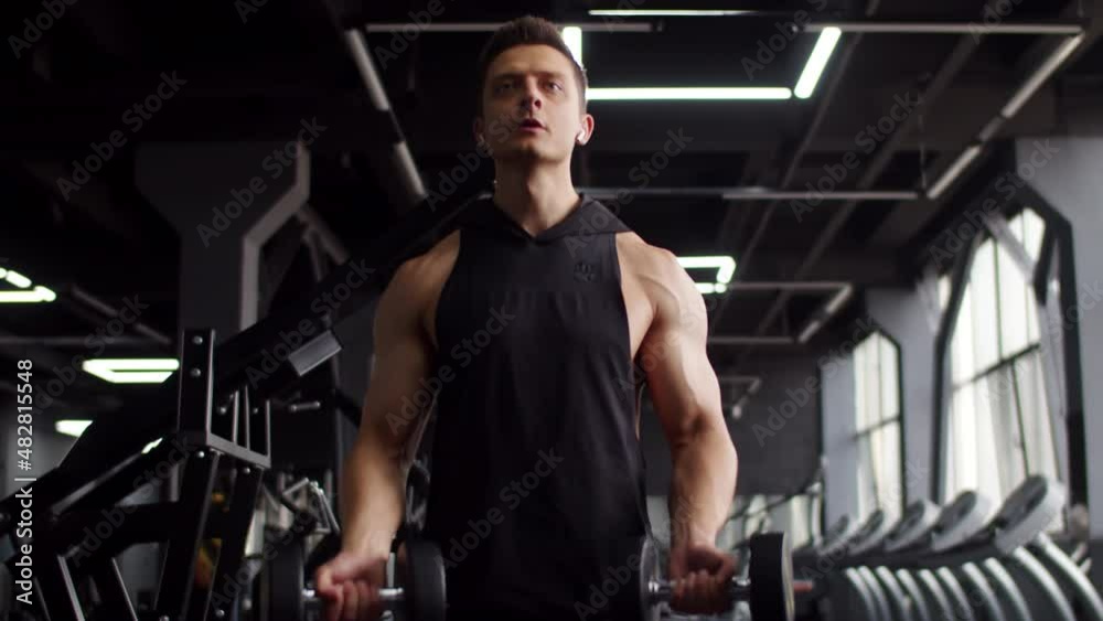 Young muscular man trains in gym lifting dumbbells with two hands, front view. Man of athletic build workout in gym with two dumbbells at hands Young muscular man trains in gym lifting dumbbells with two hands, front view. Man of athletic build workout in gym with two dumbbells at hands