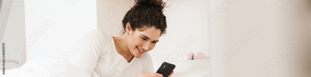 The smiling woman looking at the phone while lying on the bed in the room The smiling woman looking at the phone while lying on the bed in the room