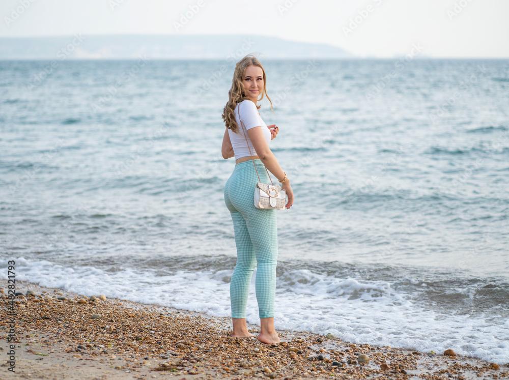 Rear view of smiling blonde woman in light blue and white standing on beach near sea.