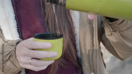 A traveler girl pours tea from a thermos cup during a hike. the girl spills a hot drink into a mug from a green thermos.