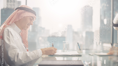 Two Successful Emirati Businessmen in White Traditional Outfit Sitting in Office and Signing Contract. Business Partners Sing Lucrative Investment. Saudi, Emirati, Arab Businessman Concept.