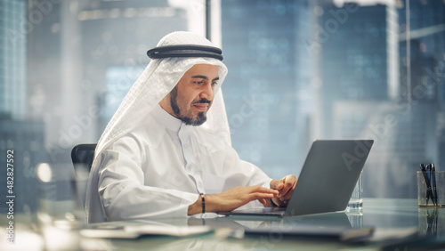 Arab Businessman in White Traditional Outfit Sitting in Office and Working on Laptop Computer. Business Manager Make Successful Investment Deal. Saudi, Emirati, Arab Businessman Concept. photo