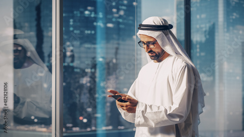 Foto Successful Muslim Businessman in Traditional White Outfit Standing in His Modern Office, Using Smartphone Next to Window with Skyscrapers