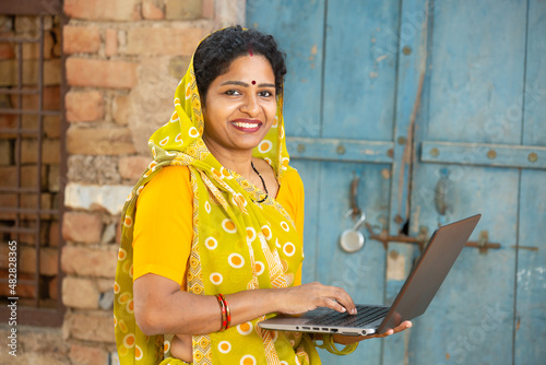 Portrait of Happy young rural indian woman using laptop. Smiling female wearing sari holding computer. She is looking at camera, skill india concept.