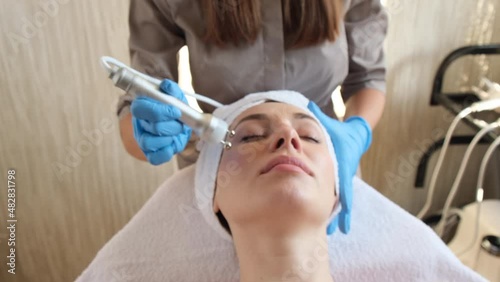 Young woman lying on cosmetologist's table during rejuvenation procedure. Cosmetologist take care about neck and face skin youthfull and wellness. Hardware face cleaning procedure   photo