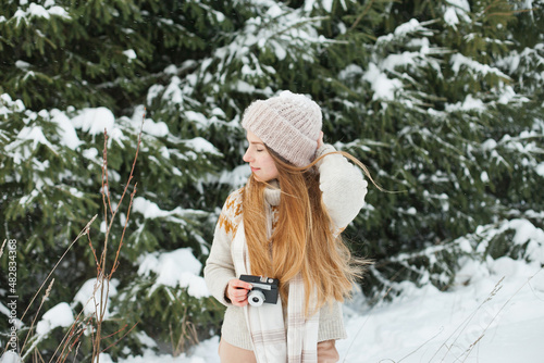 Beautiful young woman holding a retro vintage film camera for taking photos in winter snowy forest. Hobby and leisure activity outside. Hipster stylish girl in beanie hat in winter outdoors near fir.