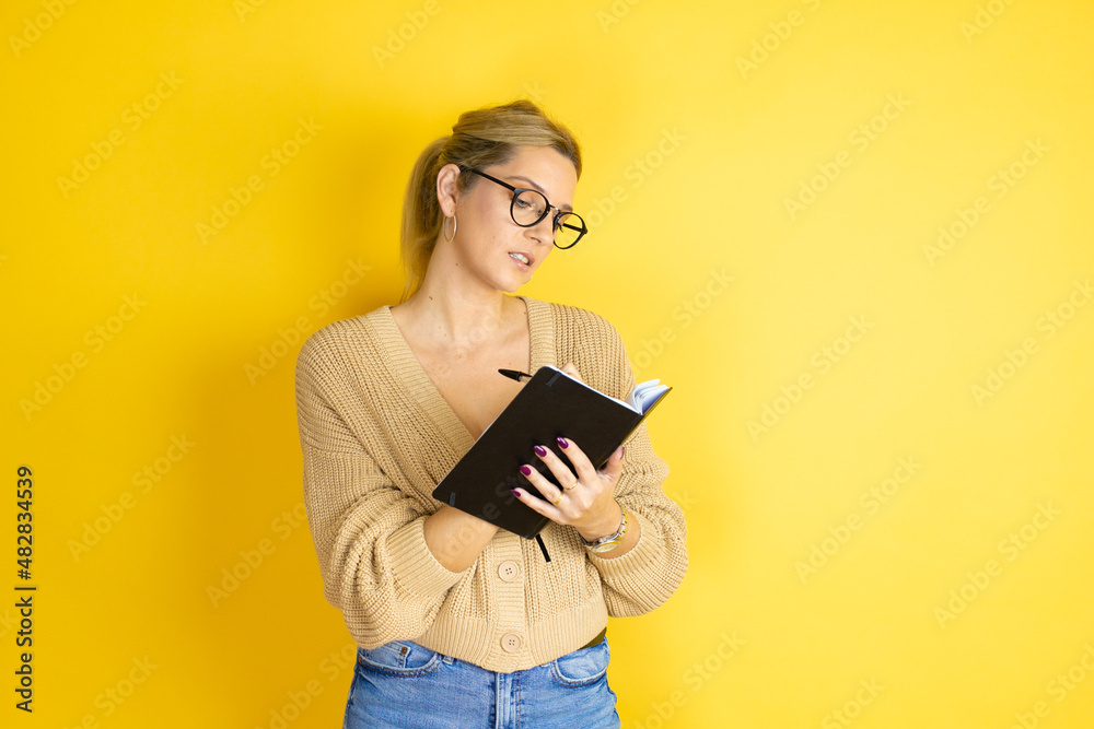 Young beautiful woman wearing casual sweater with a book in hand over isolated yellow background thinking and writing in the book Young beautiful woman wearing casual sweater with a book in hand over isolated yellow background thinking and writing in the book