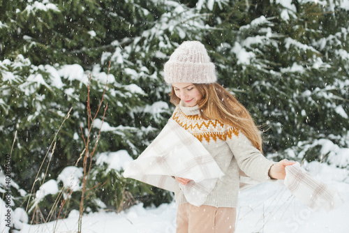 Happy woman in winter forest during snowfall, enjoying winter. Millennial young woman with long hair in beige warm outfit walking in the snowy park in nature near fir trees, being careless and free..