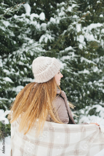 Happy woman in winter forest during snowfall, enjoying winter. Millennial young woman with long hair in beige warm outfit walking in the snowy park in nature near fir trees, being careless and free..