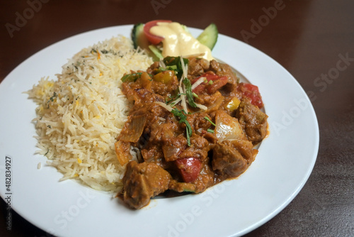 Indian curry made from meat and vegetables in a spicy sauce served with rice and salad decoration on a white plate and a brown wooden table, selected focus