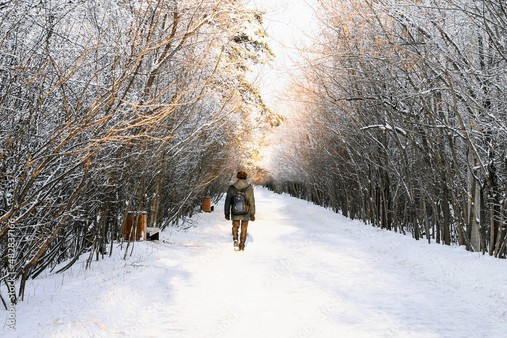 Man with a backpack walks along a wide winter road lit by the bright sun. Walk in the winter park. Beautiful bright winter landscape. Place for text. Winter season concept.