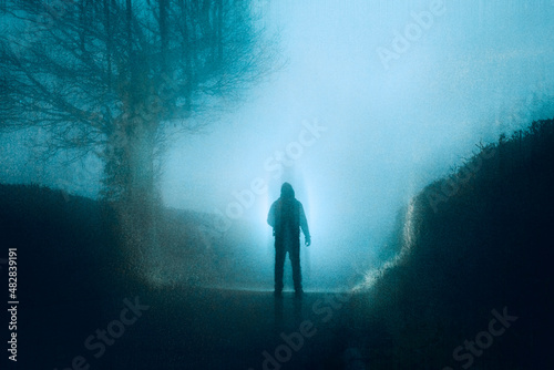 A spooky concept of a figure shining a torch on a country road. On a moody, scary foggy winters night. With a grunge, grainy edit
