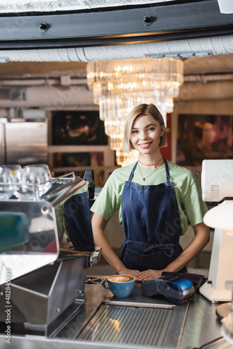 Smiling barista looking at camera near coffee and payment terminals in cafe.