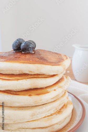 Macro close up of stack of pancakes with blueberries on top and maple syrup or honey running down the sides. Pouring jug in background set against a light airy white backdrop.
