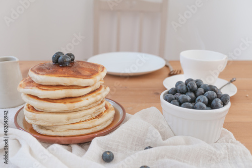Breakfast pancakes scene with no one in the shot. Hot steaming drink, blueberries and kitchen ware arranged on wood table. Rustic light and airy feel with white background and copy space available.