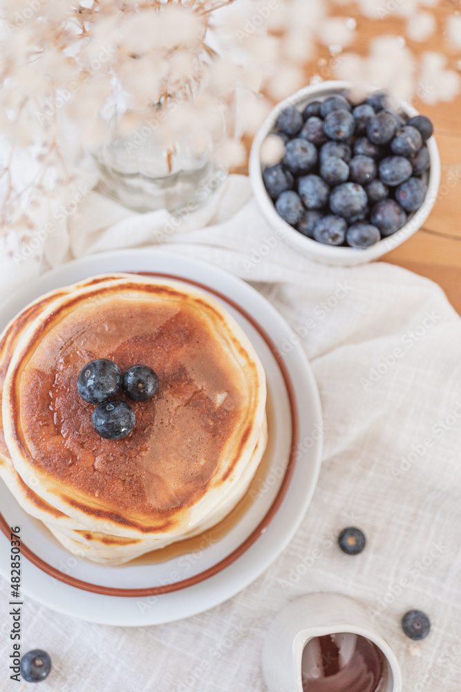 Flat lay image of a stack of pancakes with blueberries on top and maple syrup or honey running down the sides. Fruit, flowers and jug on top a wooden table. Copy space available in light airy backdrop Flat lay image of a stack of pancakes with blueberries on top and maple syrup or honey running down the sides. Fruit, flowers and jug on top a wooden table. Copy space available in light airy backdrop