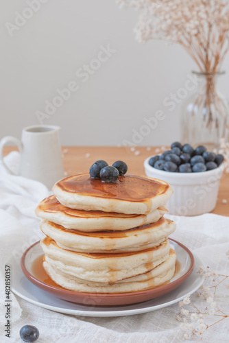 Vertical image of a stack of pancakes with blueberries on top and maple syrup or honey running down the sides. Fruit, flowers and jug on top a wooden table. Copy space available in light airy backdrop