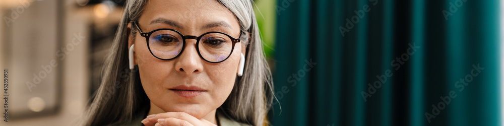 Mature grey woman in earphone working with laptop at cafe indoors Mature grey woman in earphone working with laptop at cafe indoors