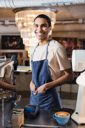 Smiling african american barista looking at camera near payment terminals and coffee in cafe. © LIGHTFIELD STUDIOS