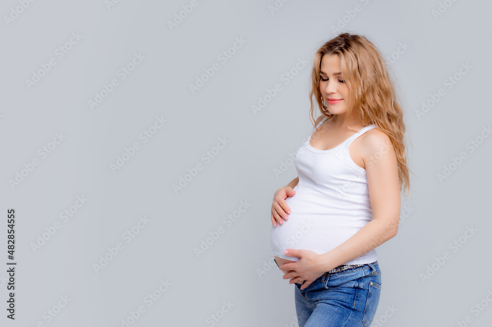 Concept Of A Happy Pregnancy. The Expectant Mother Touches Her Stomach, Smiling At The Camera Standing On A White Background. Studio shot Concept Of A Happy Pregnancy. The Expectant Mother Touches Her Stomach, Smiling At The Camera Standing On A White Background. Studio shot