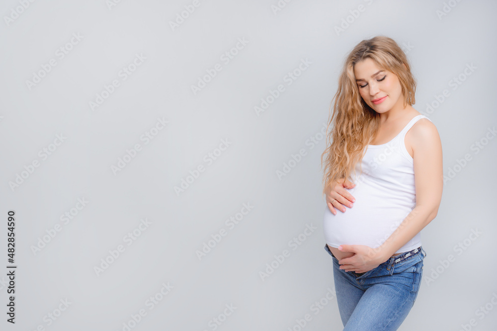 Concept Of A Happy Pregnancy. The Expectant Mother Touches Her Stomach, Smiling At The Camera Standing On A White Background. Studio shot Concept Of A Happy Pregnancy. The Expectant Mother Touches Her Stomach, Smiling At The Camera Standing On A White Background. Studio shot