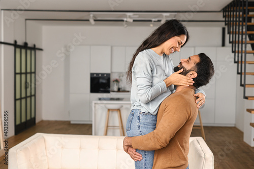 Cheerful Indian guy holding happy woman in arms while standing in apartment. Ethnic latin hispanic couple in love relocating in own flat. Move in new house concept