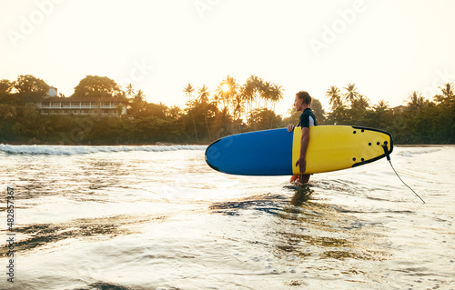 Teen boy with blue and yellow surfboard entering the waves for surfing with sunset rays on palm trees grove. Happy childhood and active sporty people vacation time concept.