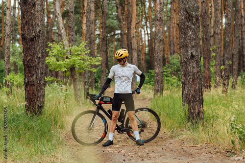 Male cyclist in outfit stands with a bicycle in the woods on a trail on a background of pine forest and poses for the camera.