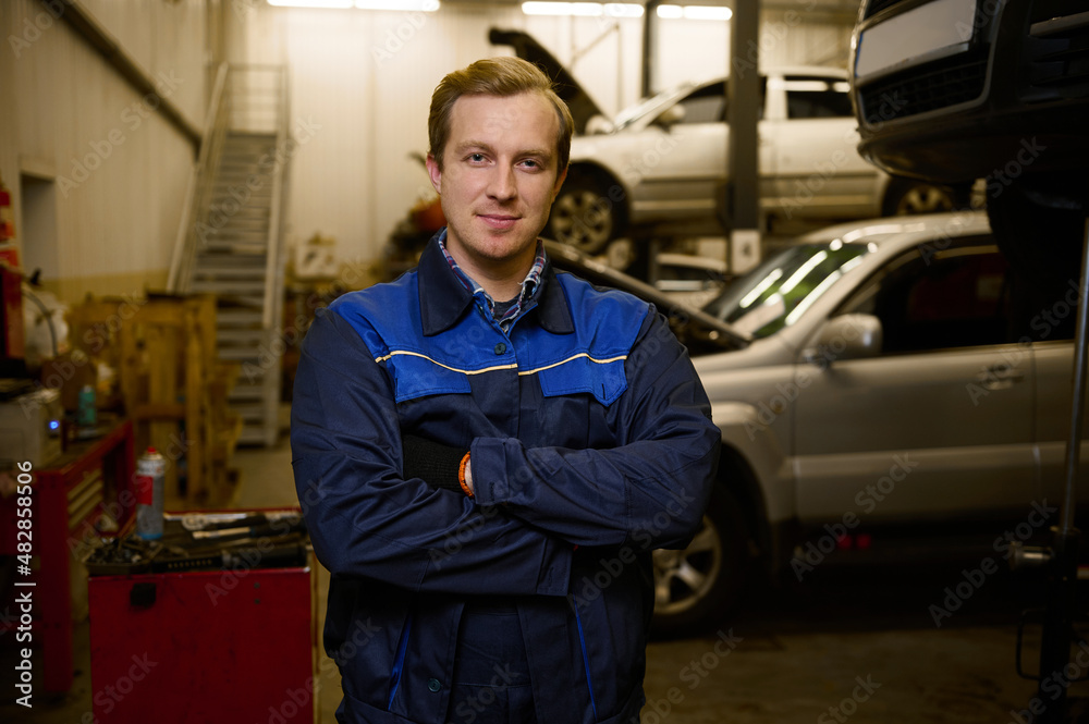 Confident portrait of an auto mechanic dressed in professional uniform looking at camera standing with crossed arms in car repair shop. Car repair, diagnostics and professional service in car service Confident portrait of an auto mechanic dressed in professional uniform looking at camera standing with crossed arms in car repair shop. Car repair, diagnostics and professional service in car service