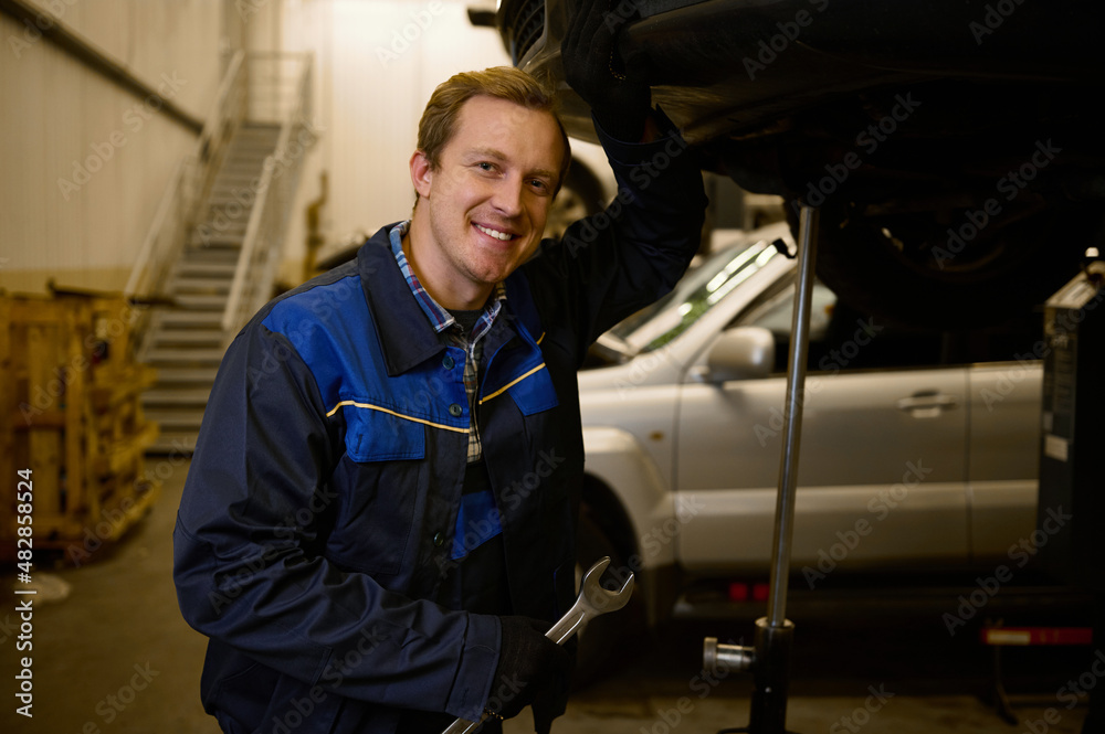 Portrait of a young mechanic in professional uniform, holding a wrench and working in auto service with lifted vehicle, smiles toothy smile lookng at camera. Car repair and maintenance. Portrait of a young mechanic in professional uniform, holding a wrench and working in auto service with lifted vehicle, smiles toothy smile lookng at camera. Car repair and maintenance.
