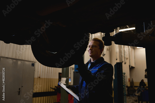 Auto mechanic in his workshop looking under a car on a hoist. Car maintenance and warranty repair concept in repair shop garage. Car service