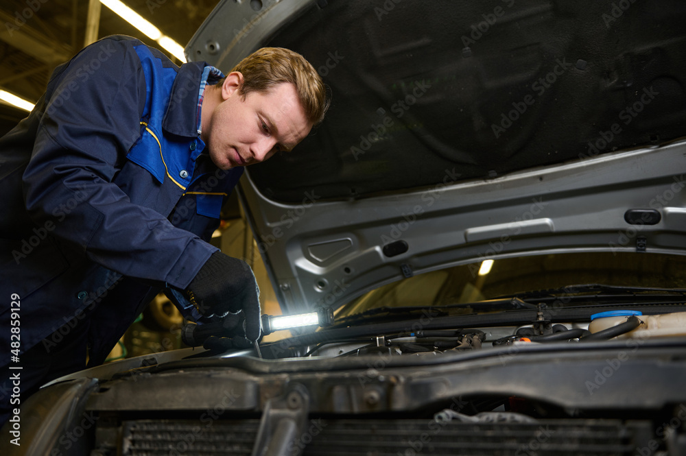 Technician, garage mechanic using flashlight luminous lamp while inspecting a car under hood in repair shop. Auto service, car maintenance concept Technician, garage mechanic using flashlight luminous lamp while inspecting a car under hood in repair shop. Auto service, car maintenance concept