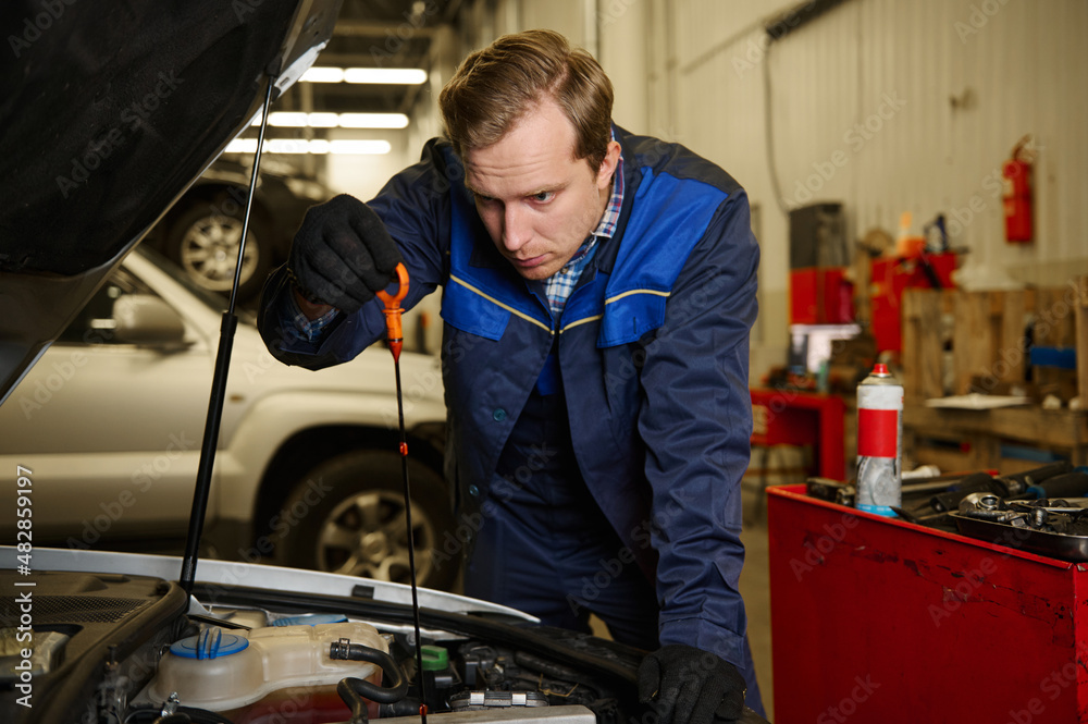 Car engineer technician mechanic in professional uniform checking oil level in car engine while doing repair and maintenance in the automobile in the repair shop garage. Car engineer technician mechanic in professional uniform checking oil level in car engine while doing repair and maintenance in the automobile in the repair shop garage.